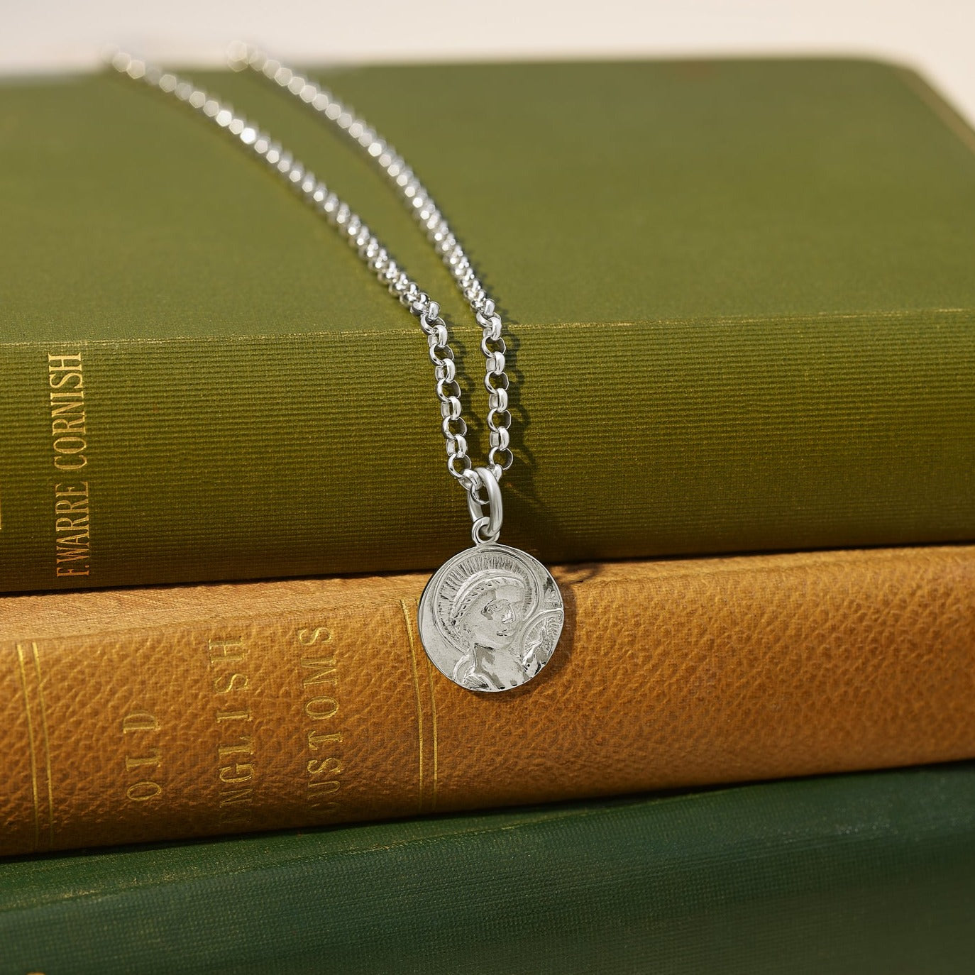 Handcrafted round sterling silver necklace pendant featuring an engraving of Saint Anthony, suspended from a sterling silver chain, resting atop two stacked books.