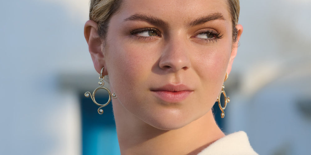 Model wears handcrafted 9ct yellow gold earring drops with a circular shape and spiral at four points, hanging from hoop earrings.
