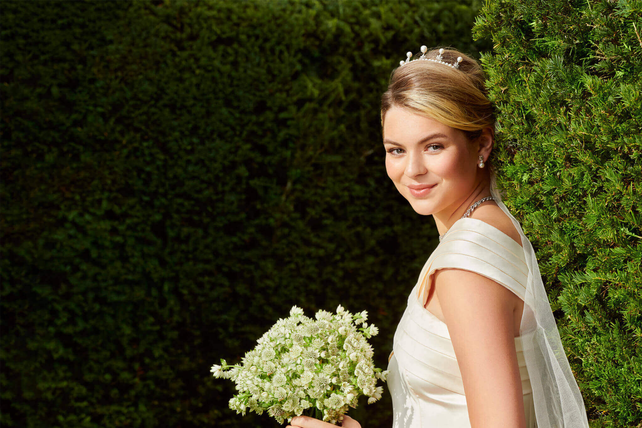 An image of a bride wearing a tiara, pearl earrings and a diamond bracelet, against a background of green bushes.