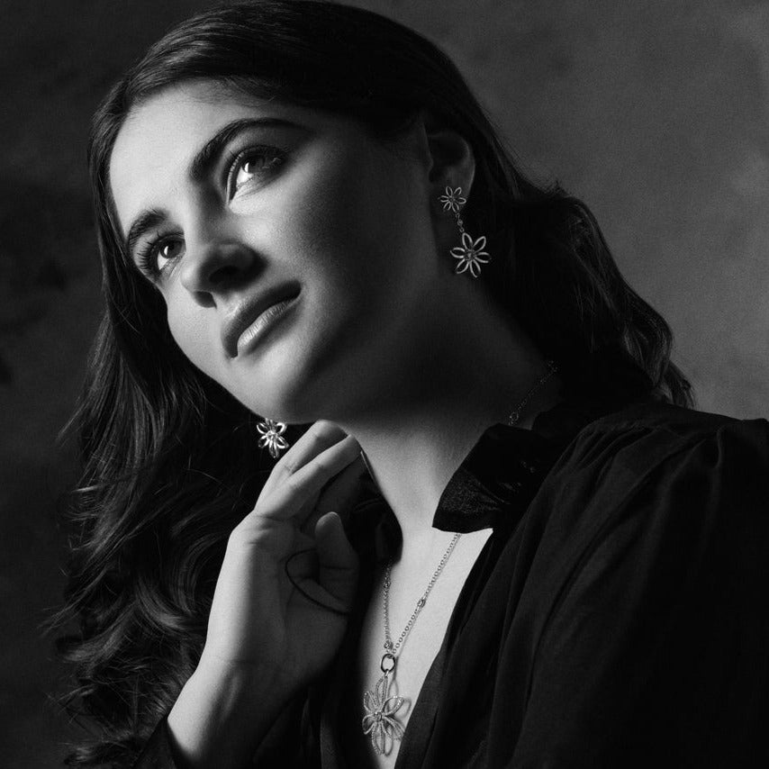 Model wearing handcrafted sterling silver earring drops with a larger flower suspended below a smaller one, each set with blue topaz, birthstone of November.