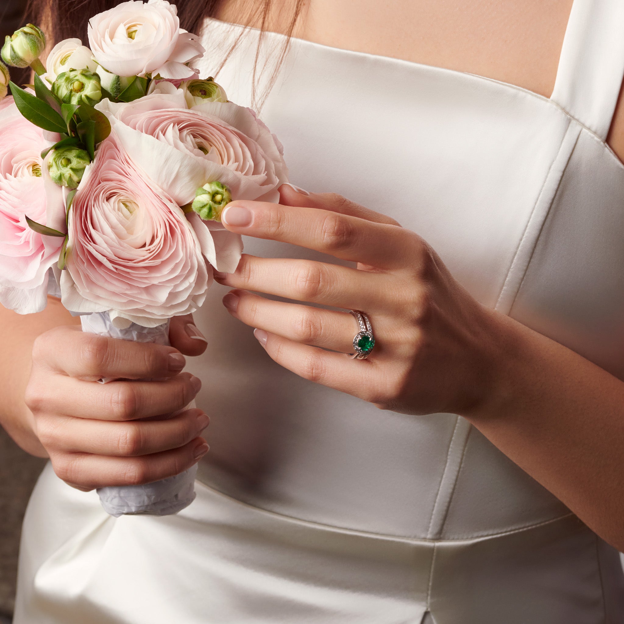 Model wearing a handcrafted 18ct white gold engagement ring with a round emerald at the centre, surrounded by smaller diamonds. The band of the engagement ring is also set with small diamonds.