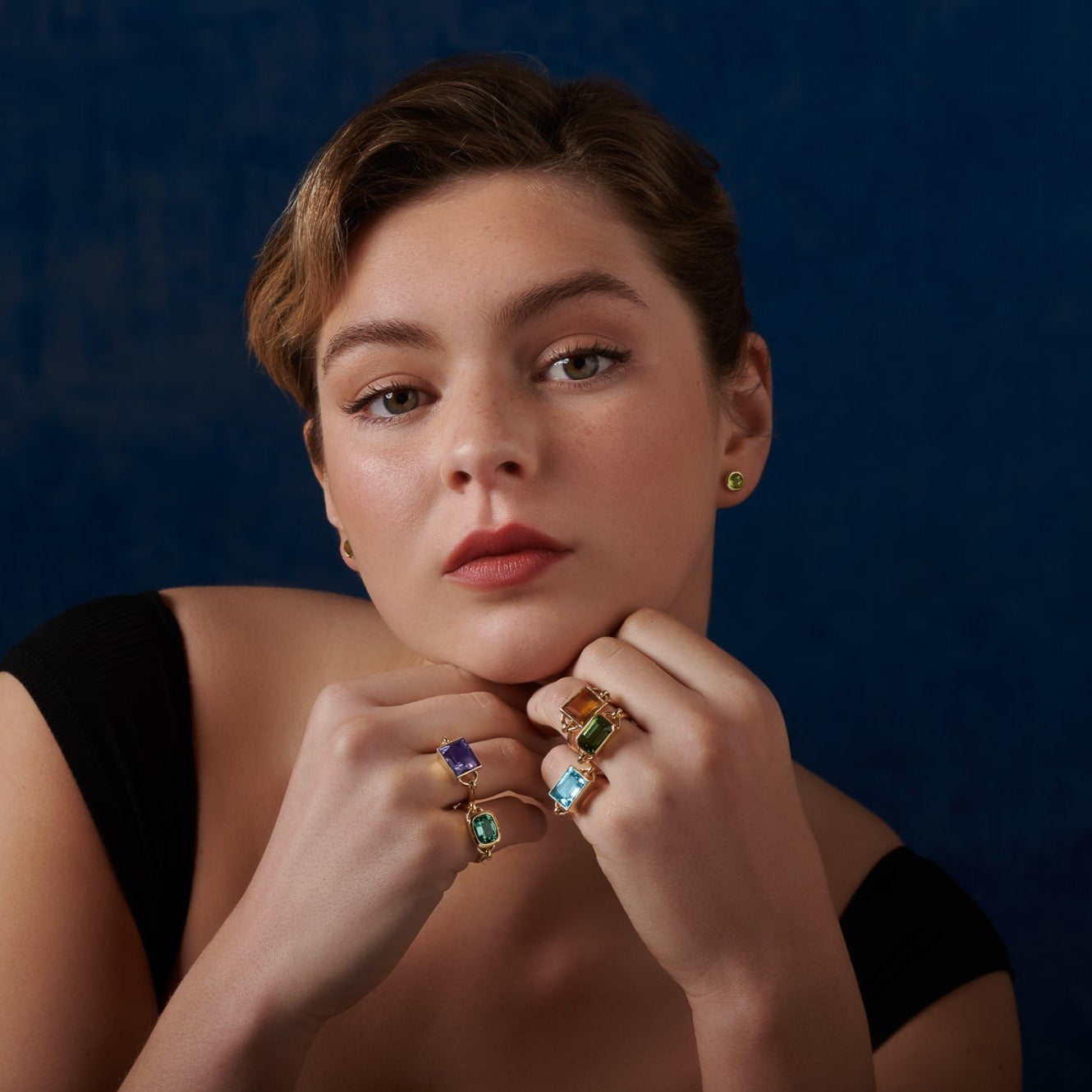 Model wearing handcrafted rings in yellow gold featuring various emerald-cut precious stones set in the centre with two small loops on either side.