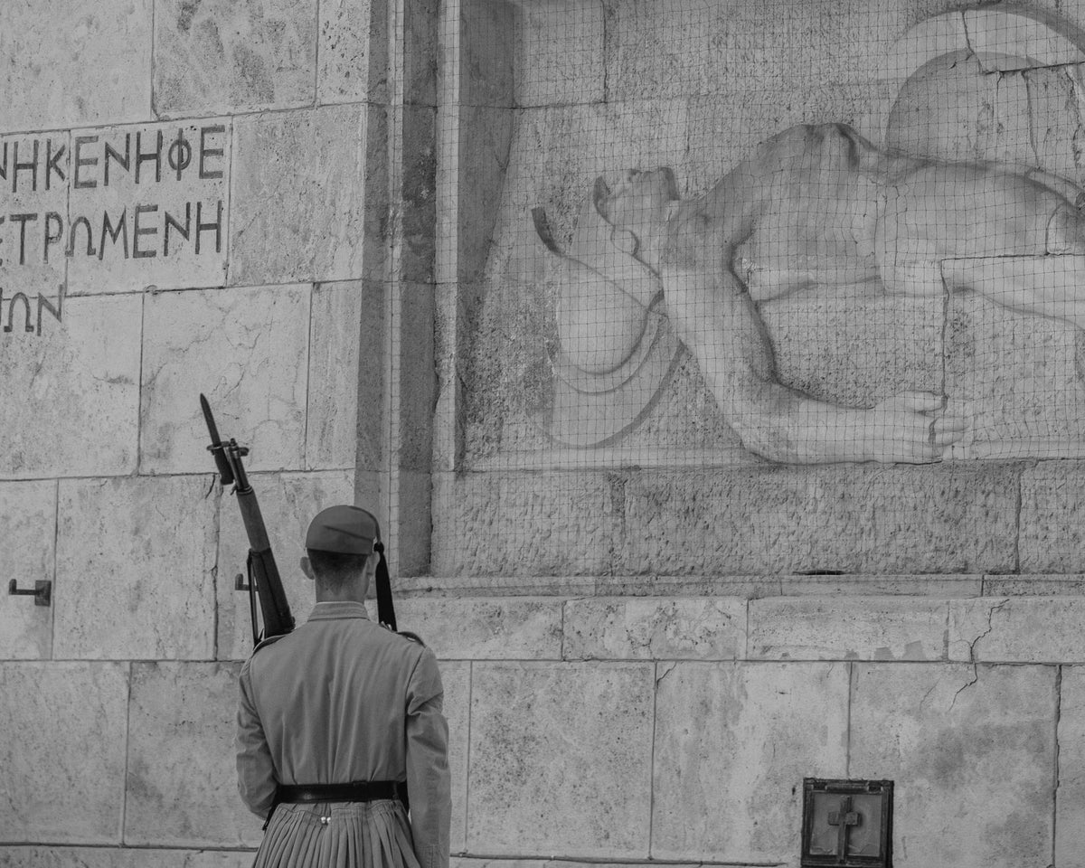 Greek soldier in black and white facing a historic stone relief