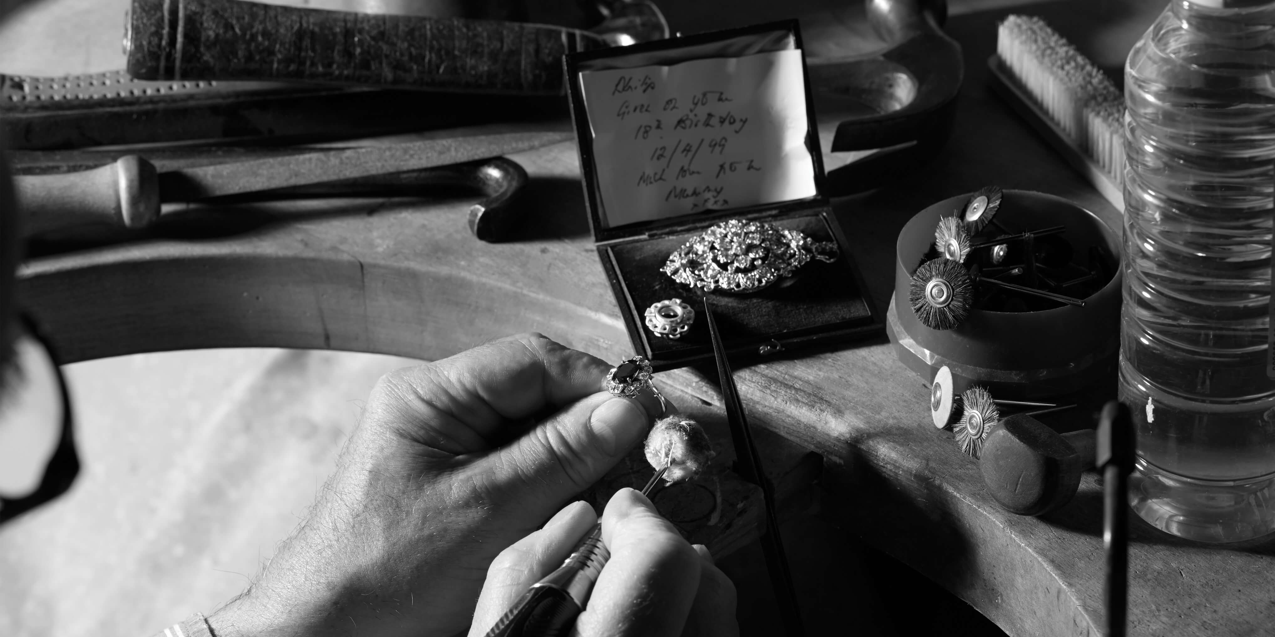 Jeweller hand-setting a gemstone in a bespoke ring at a workbench with traditional tools and an old brooch.