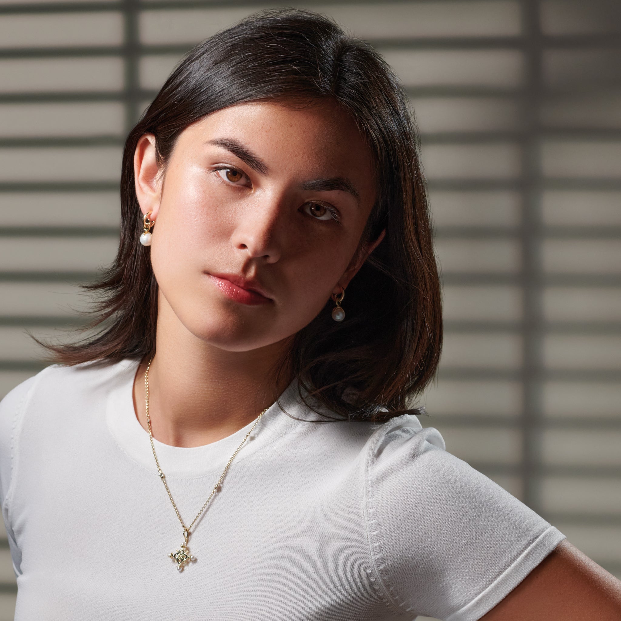 Model wearing a handcrafted cross necklace pendant in precious metal with precious stone and earring drops with round precious stones in precious metal.