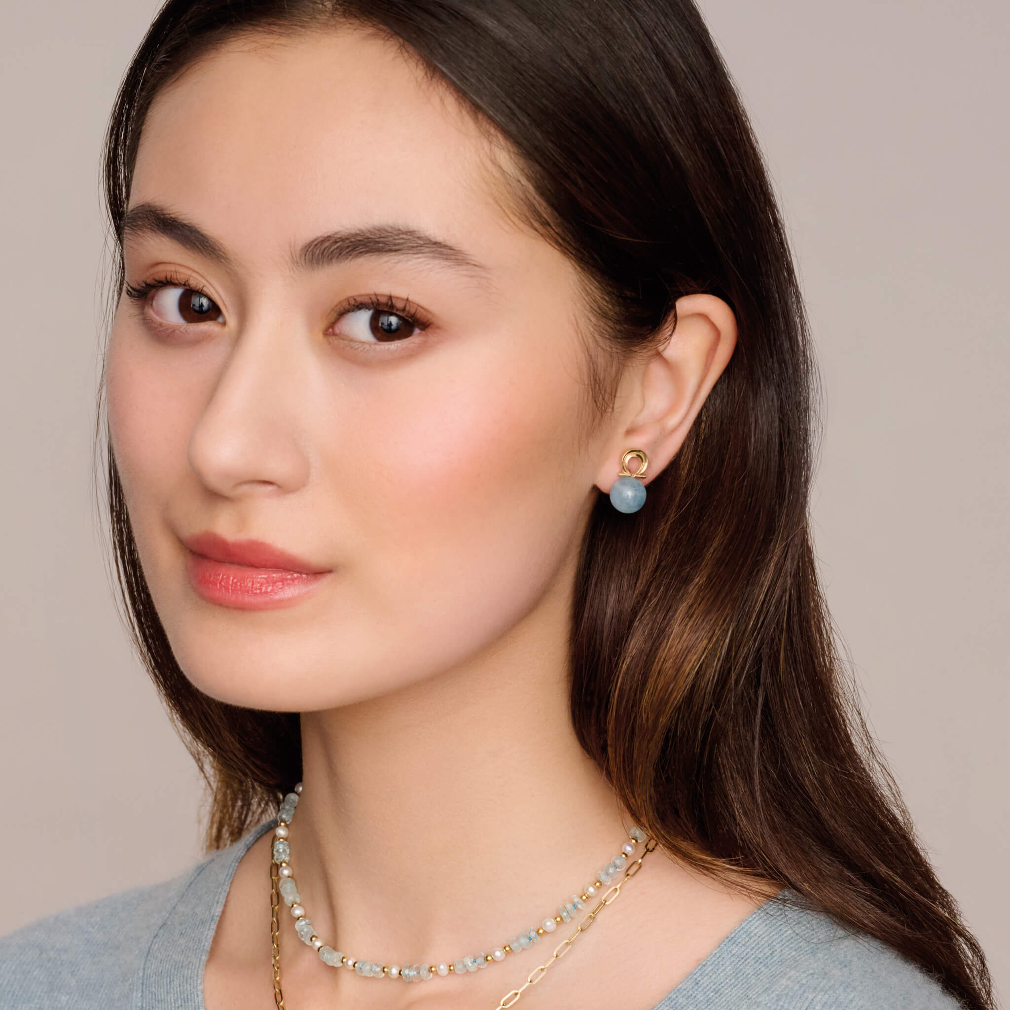 Model wears handcrafted 9ct yellow gold pair of studs with aquamarine beads. One stud is the shaped of alpha Greek letter and the other omega letter.