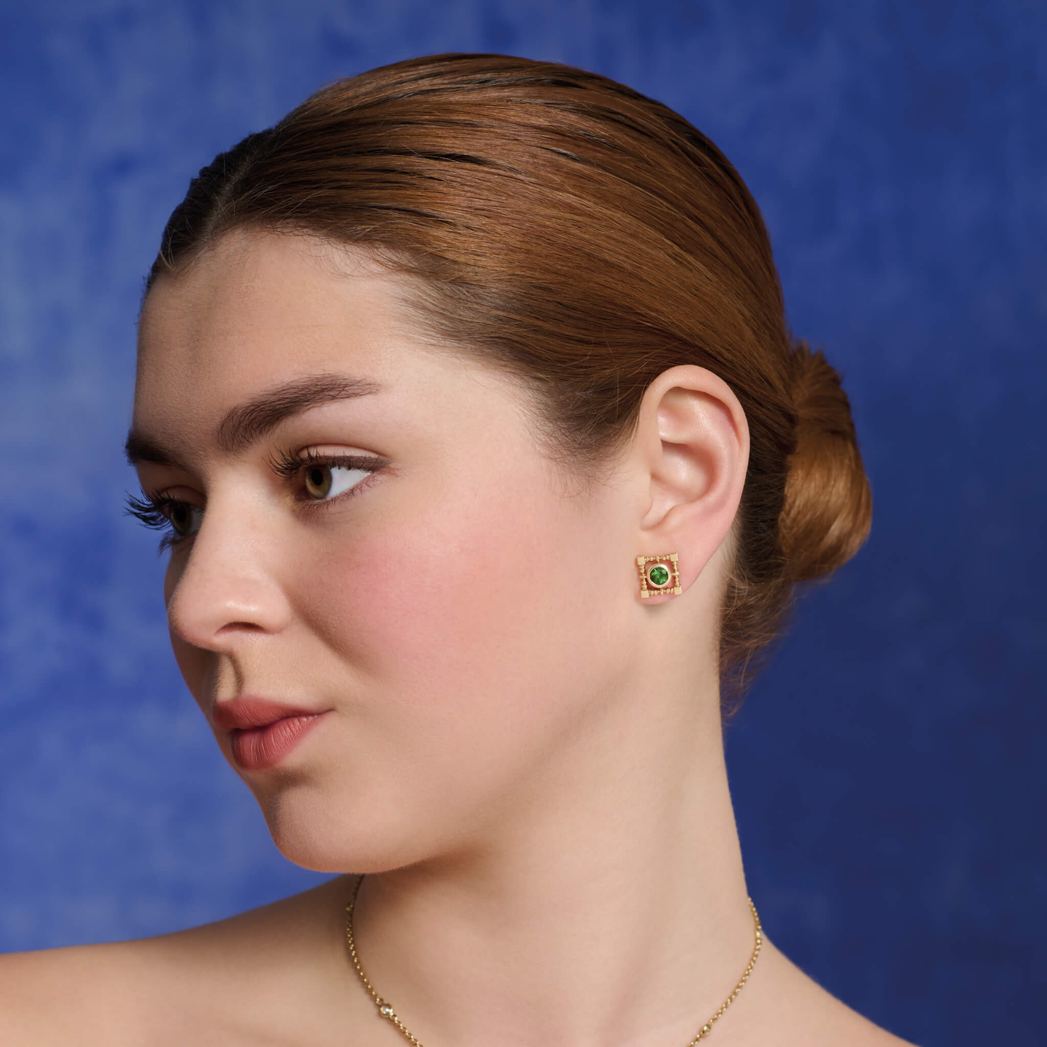 Model wears handcrafted yellow gold stud earrings in a square lattice shape with a central green tourmaline.