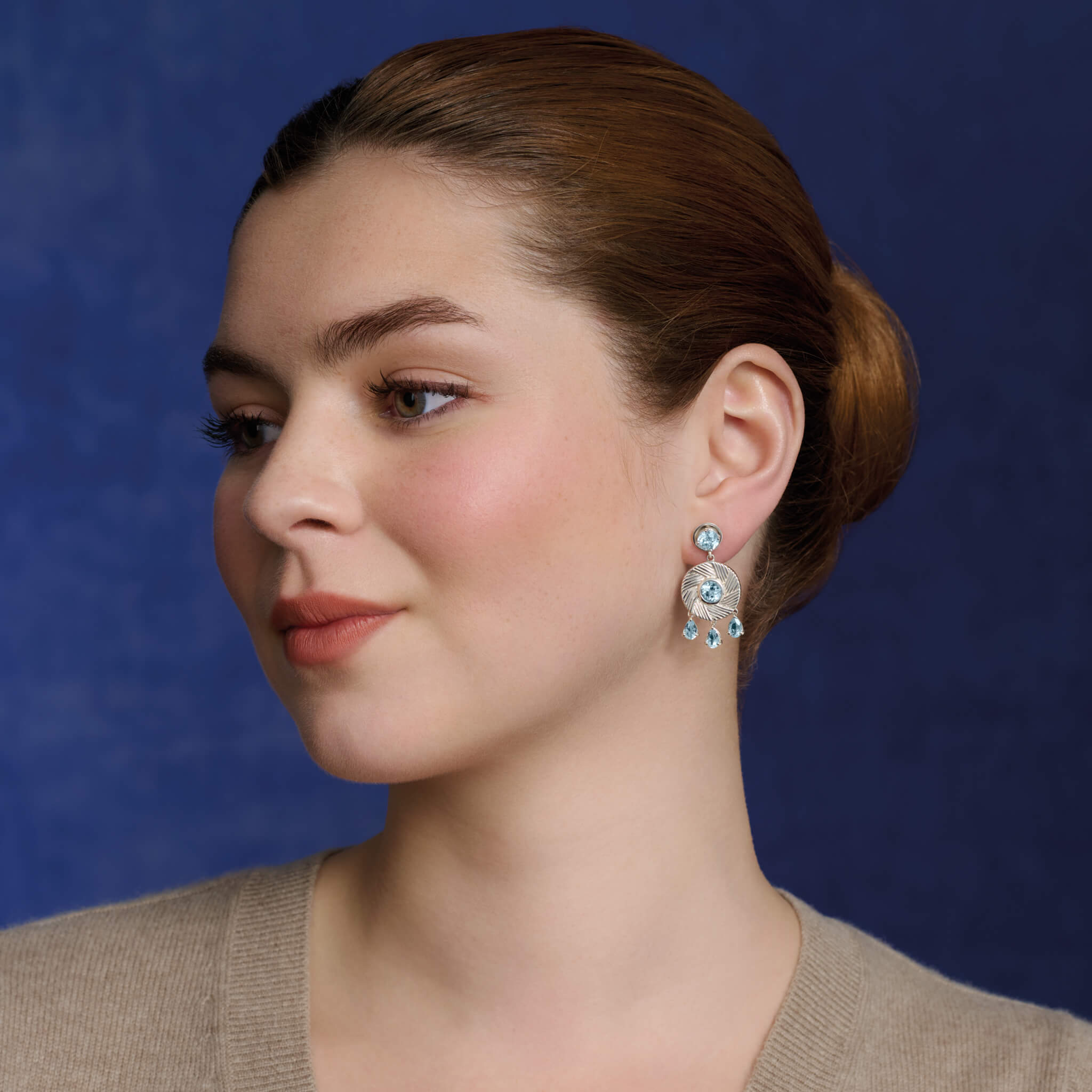 Model wears handcrafted sterling silver earrings with a round design featuring engraved stripes, centered with a blue topaz, and accented by three dangling blue topaz drops.