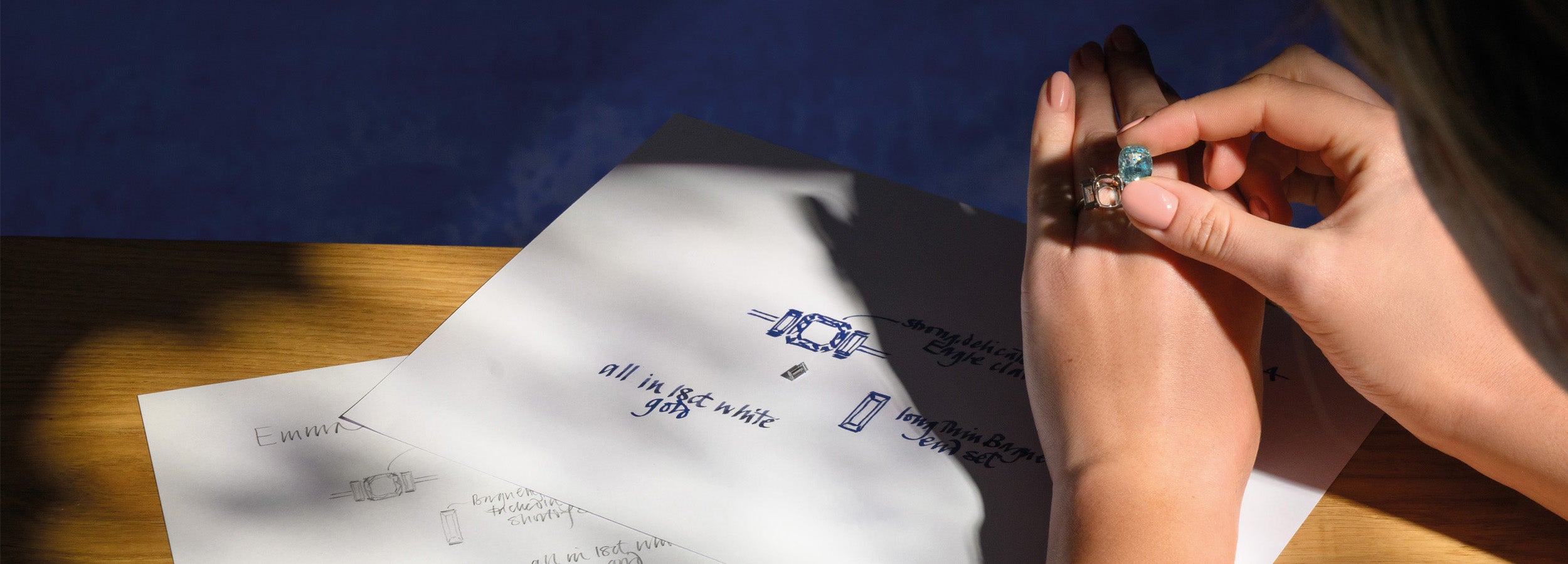 A woman planning the design of her engagement ring, holding a precious gemstone over engagement ring sketches.