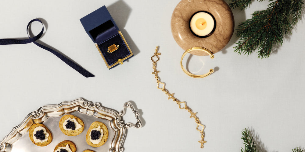 Yellow gold jewellery on white table cloth. Also on table are a ribbon, silver tray with capanes, candle and pine branches.