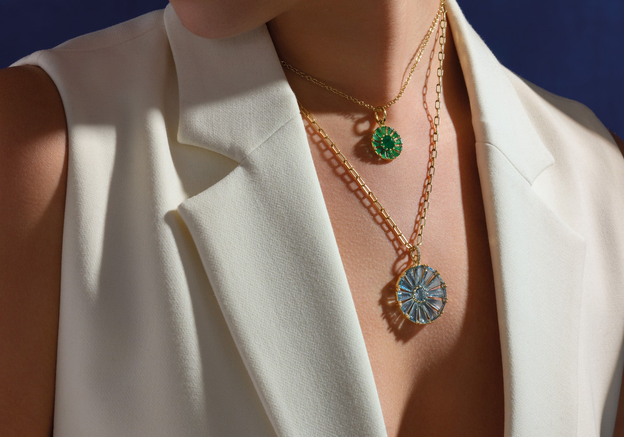 Model wearing two handcrafted circular necklace pendants featuring baguette cut emeralds and blue topaz with central precious gemstones.