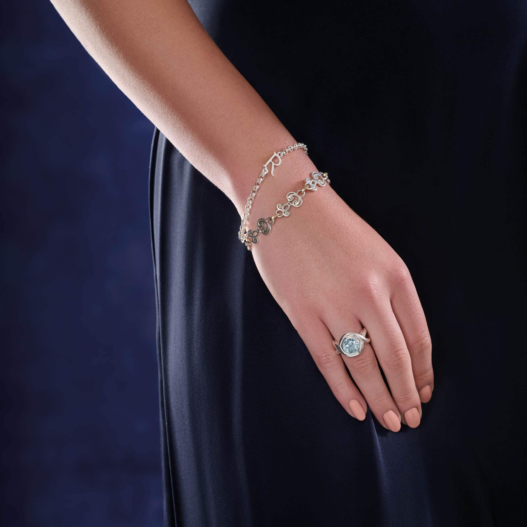 Model wearing a handcrafted ring in precious metal featuring curved shanks with a round large precious stone, and sterling silver bracelet with a central silver letter accent.