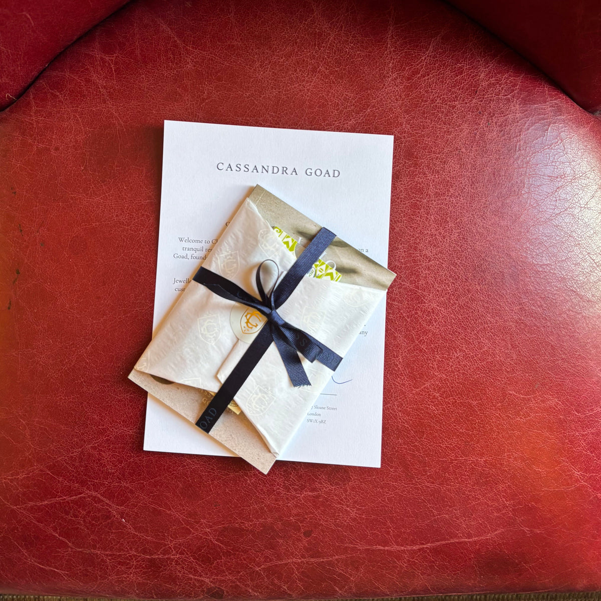 A small kraft paper envelope tied with a dark ribbon sits on top of a white card printed with the name Cassandra Goad.