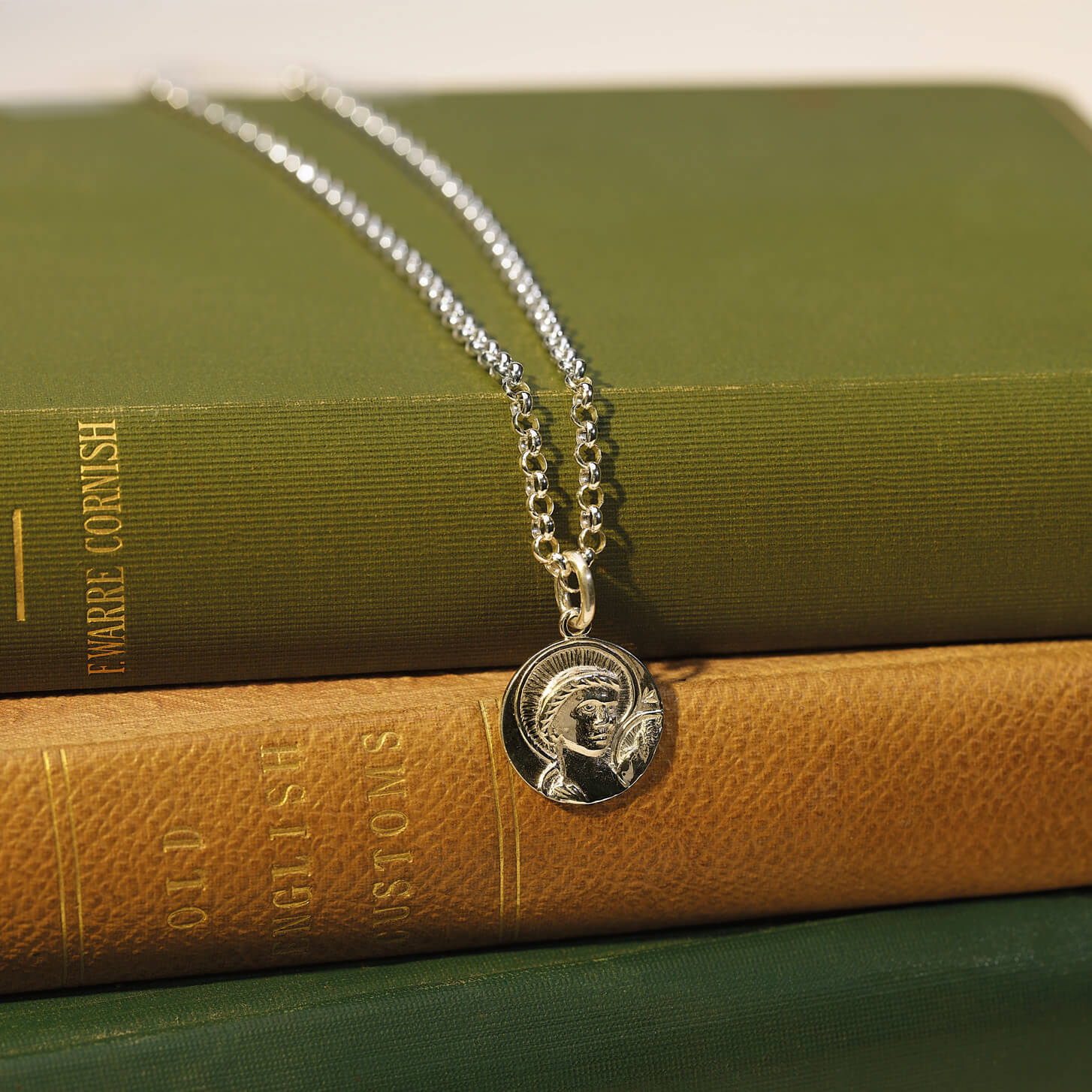 Handcrafted silver round necklace pendant engraved with a saint and a chain necklace laid on some green books.