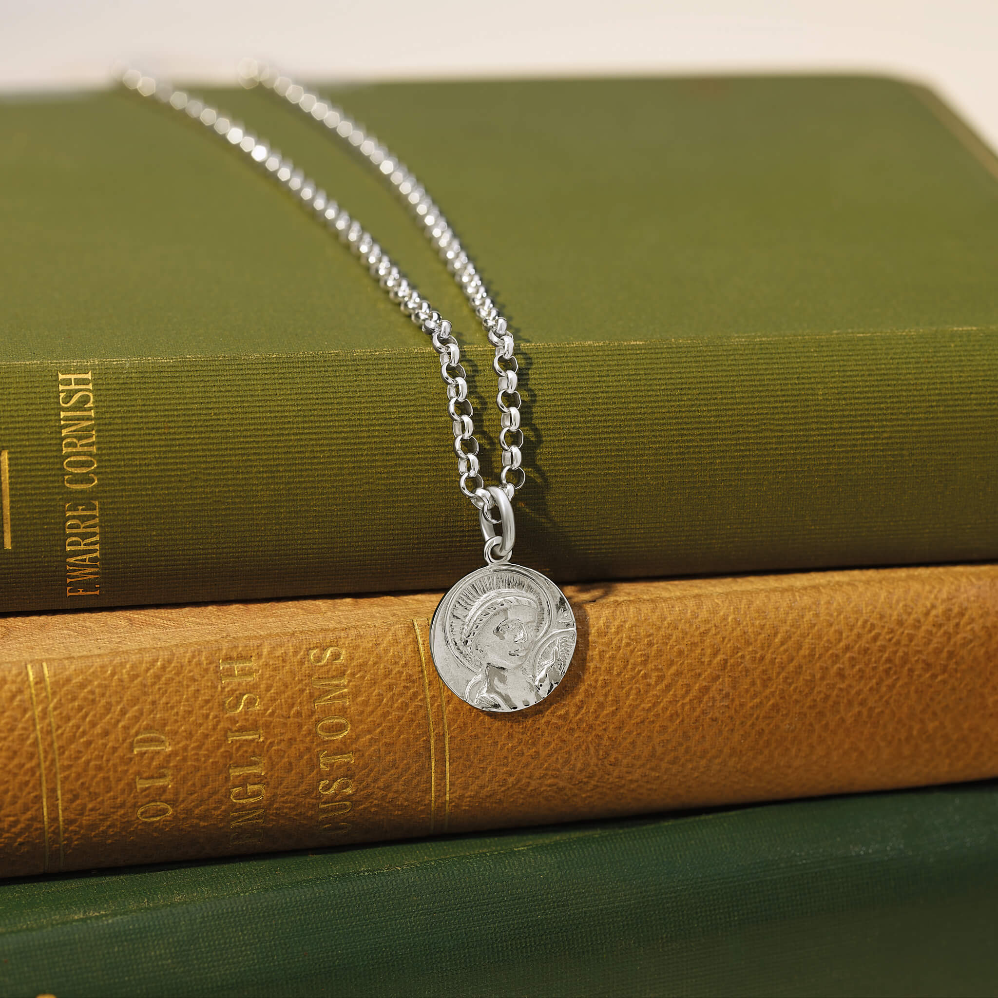 Handcrafted sterling silver round necklace pendant engraved with a saint and a chain necklace laid on some green books.