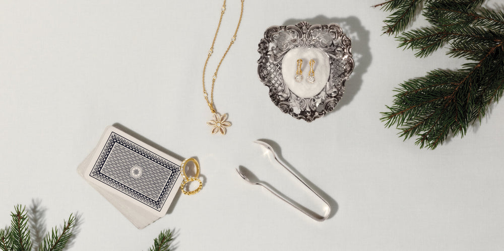 Diamond jewellery on white cloth table with navy playing cards, silver sugar tongs and spruce branches.