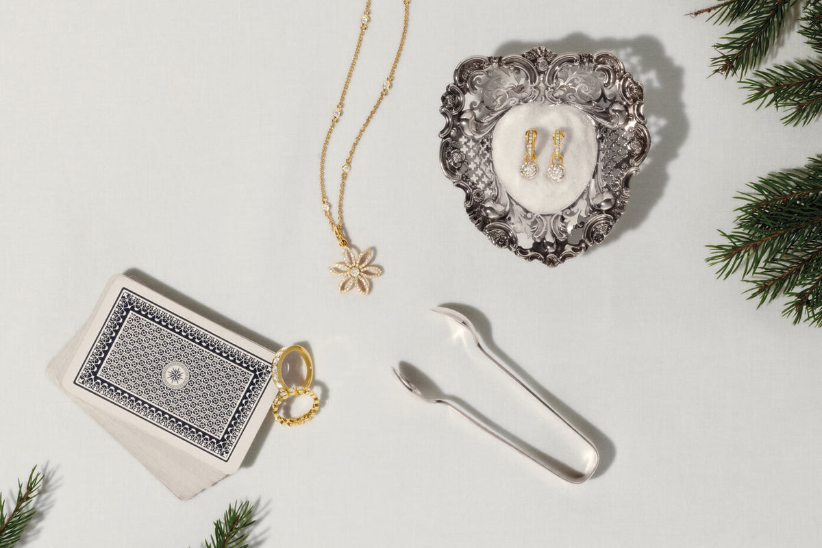 Diamond jewellery on white cloth table with navy playing cards, silver sugar tongs and spruce branches.