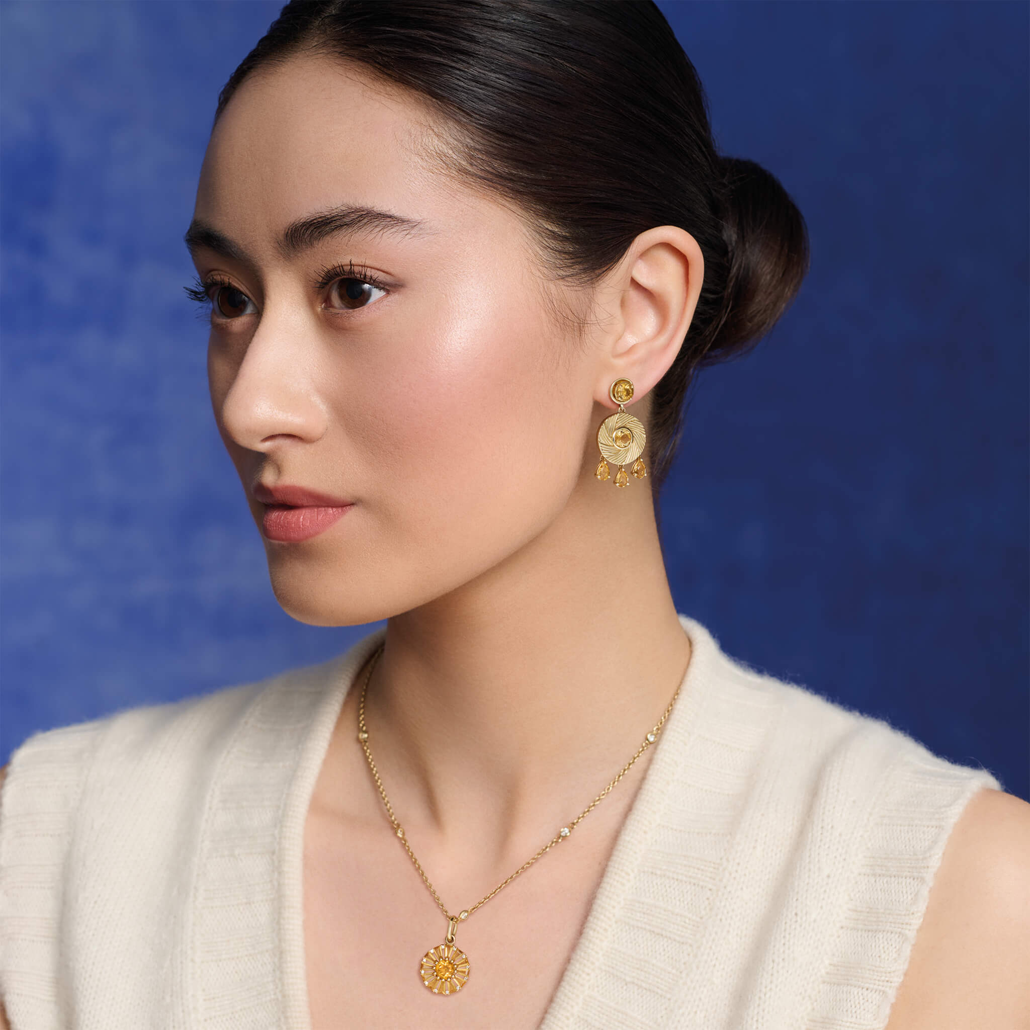 Model wears handcrafted yellow gold earrings with a round design featuring engraved stripes, centered with citrine, and accented by three dangling blue topaz drops, november birthstone