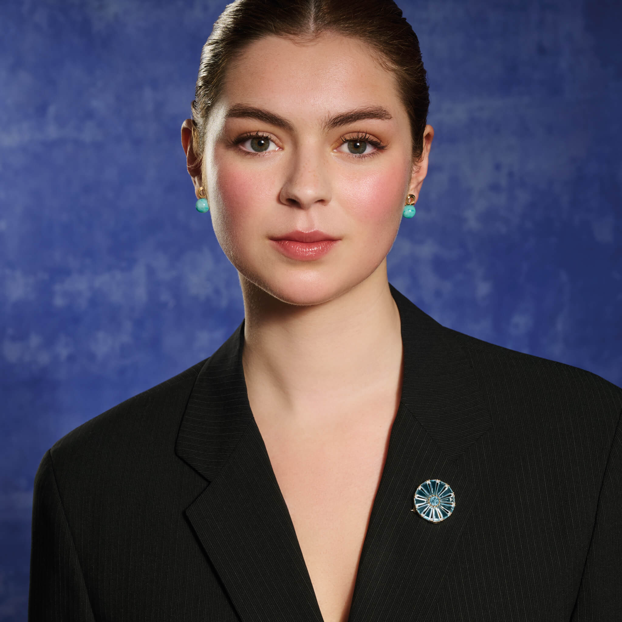 Model wears handcrafted yellow gold circular brooch with baguette cut blue topaz, a central blue topaz and diamond gemstones.