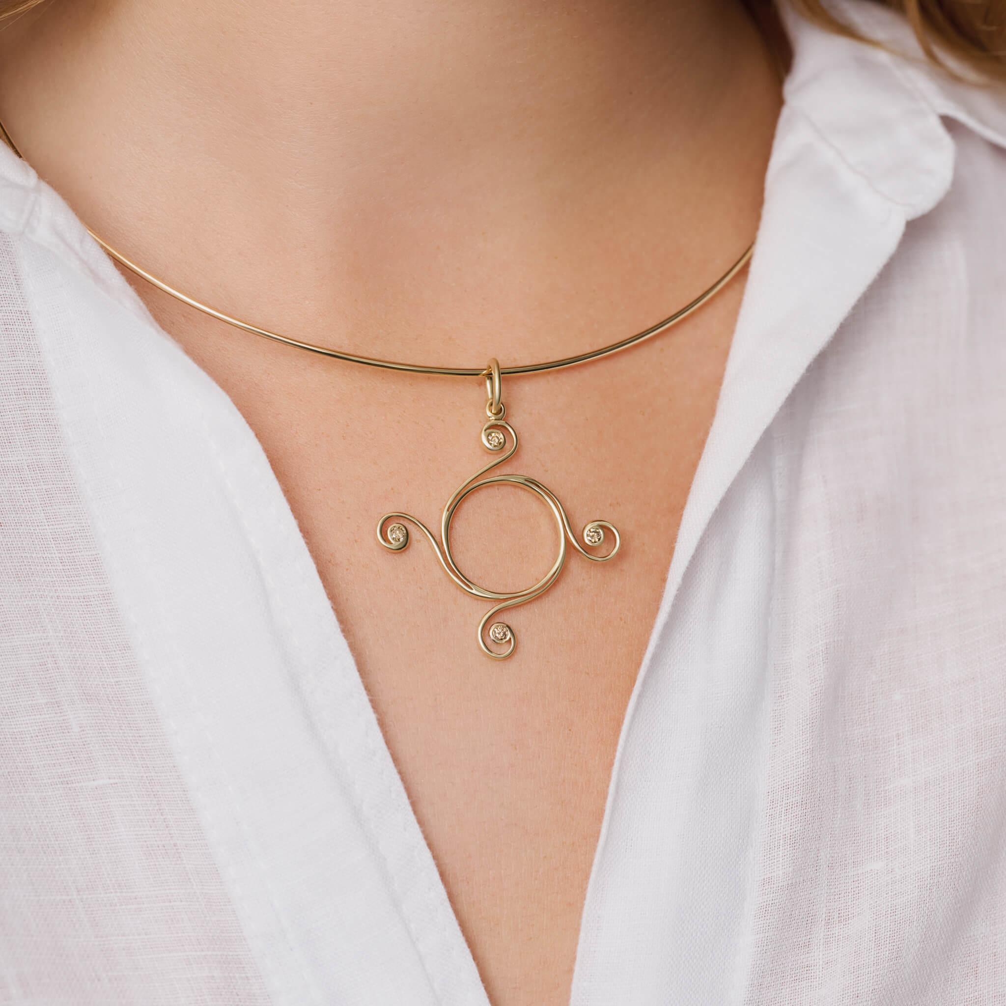 Model wears handcrafted yellow gold Athena necklace pendant with a circular shape and spiral at four points, hanging on gold chain with brown diamonds.