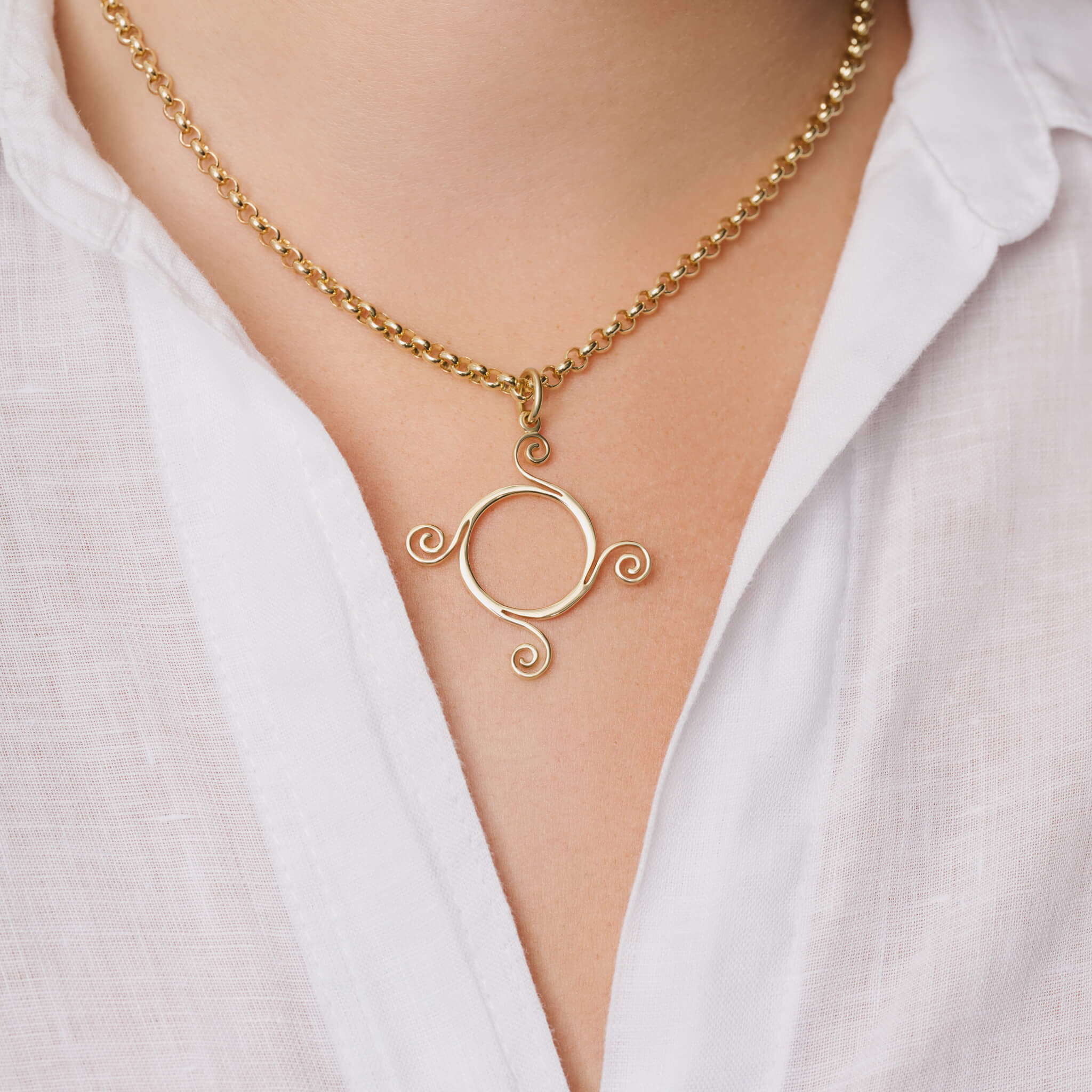 Model wears handcrafted yellow gold Athena necklace pendant with a circular shape and spiral at four points, hanging on gold chain.