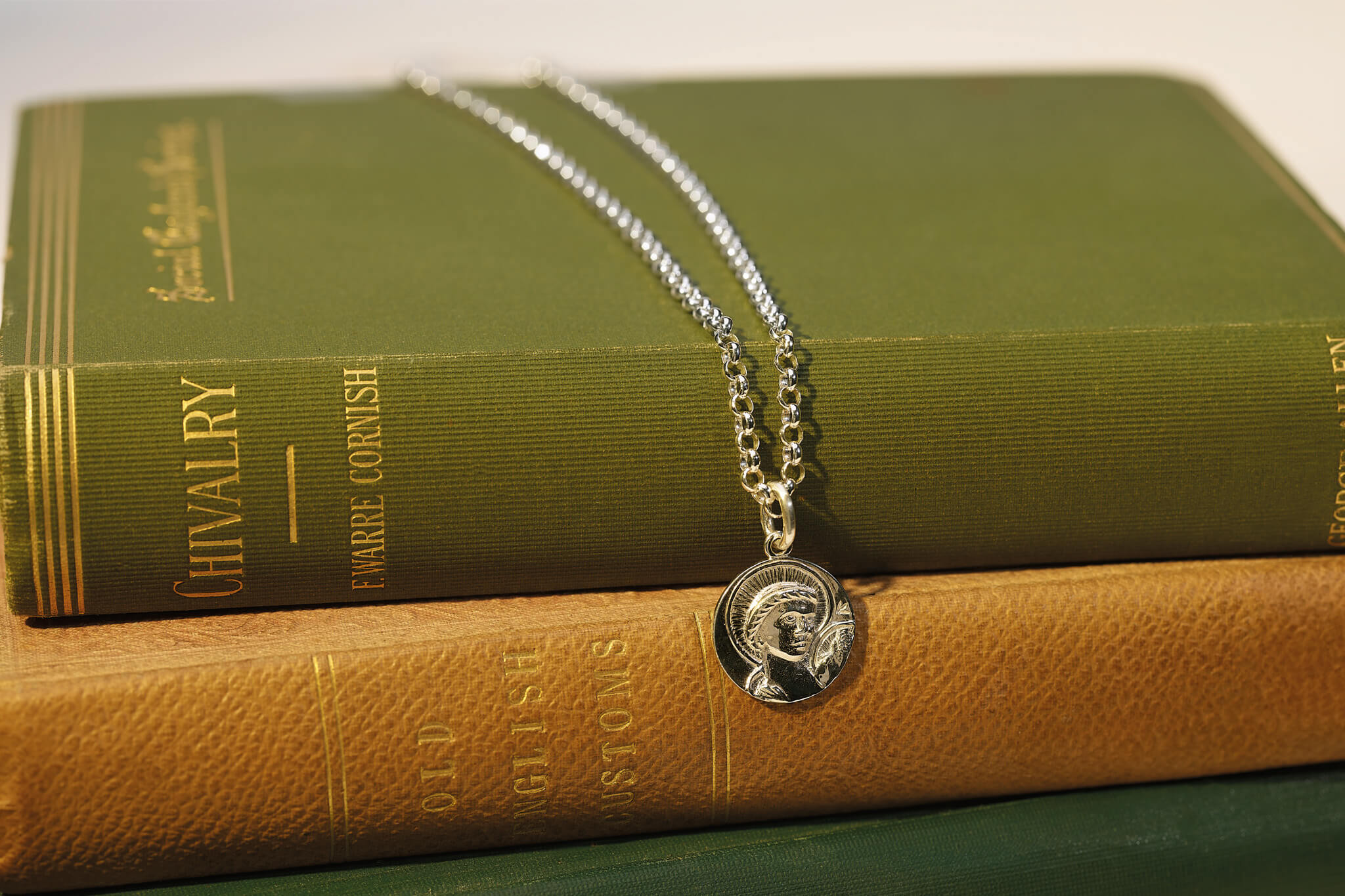 Handcrafted silver round necklace pendant engraved with a saint and a chain necklace laid on some green books.