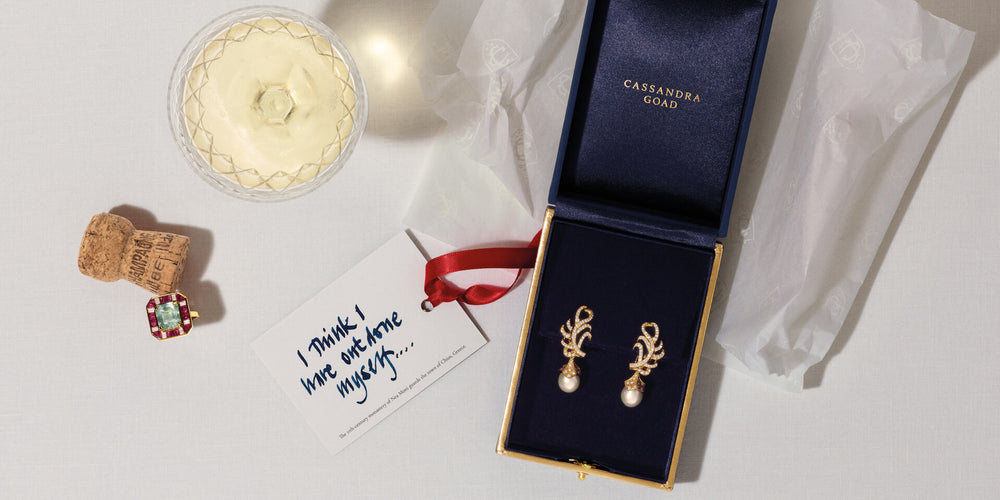 CG box containing Fouquet earring drops in yellow gold with pearl hanging on Feather Plume earrings in 18ct yellow gold. Place des Vosges ring next to wine cork. Handwritten note on table.