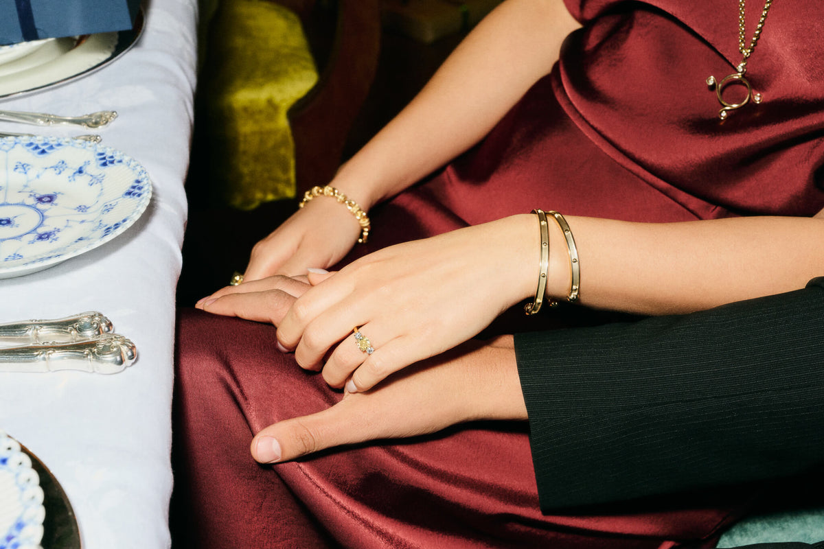 Two models embrace hands at a Christmas dinner table. The woman wears Cassandra Goad golden jewellery including 9ct gold Decimus cuffs, a golden bracelet and a yellow diamond Delilah engagement ring.