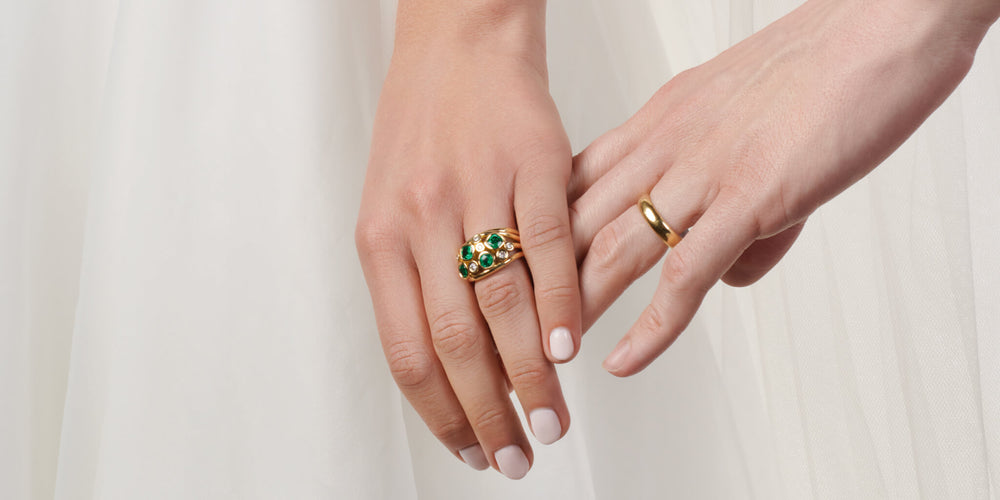 A woman's hand gently touching a man's hand, showcasing a handcrafted 18ct yellow gold engagement ring set with precious stones on hers, alongside a yellow gold wedding band on his.