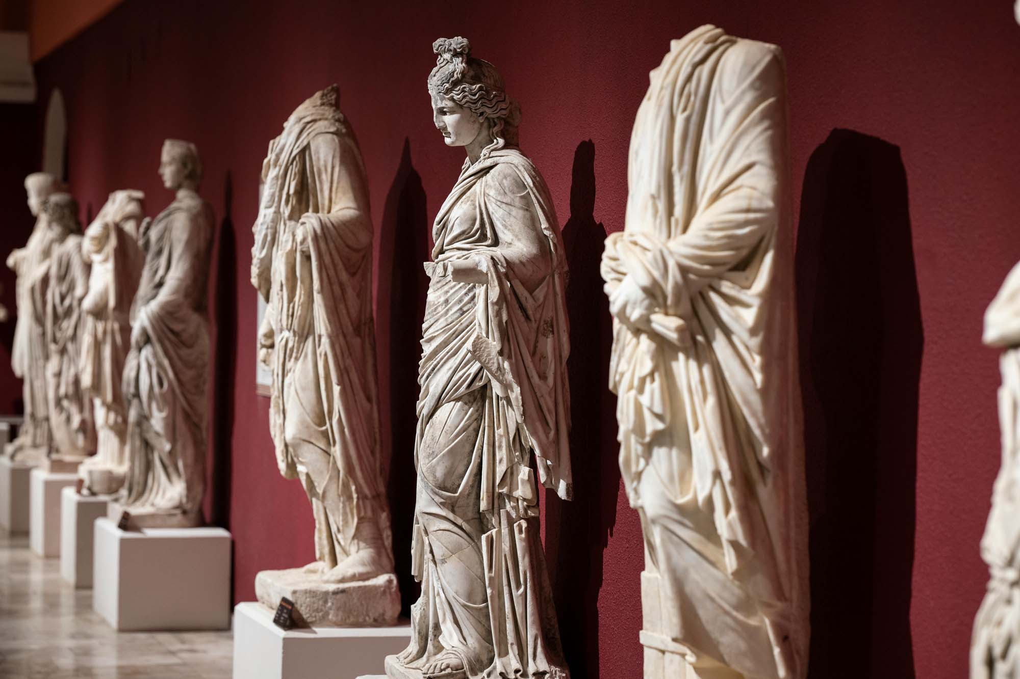 Line formed by several full body ancient Greek marble sculptures in a Museum. The seven sculptures are stood against a red painted wall making the whole of the marble stand out. 