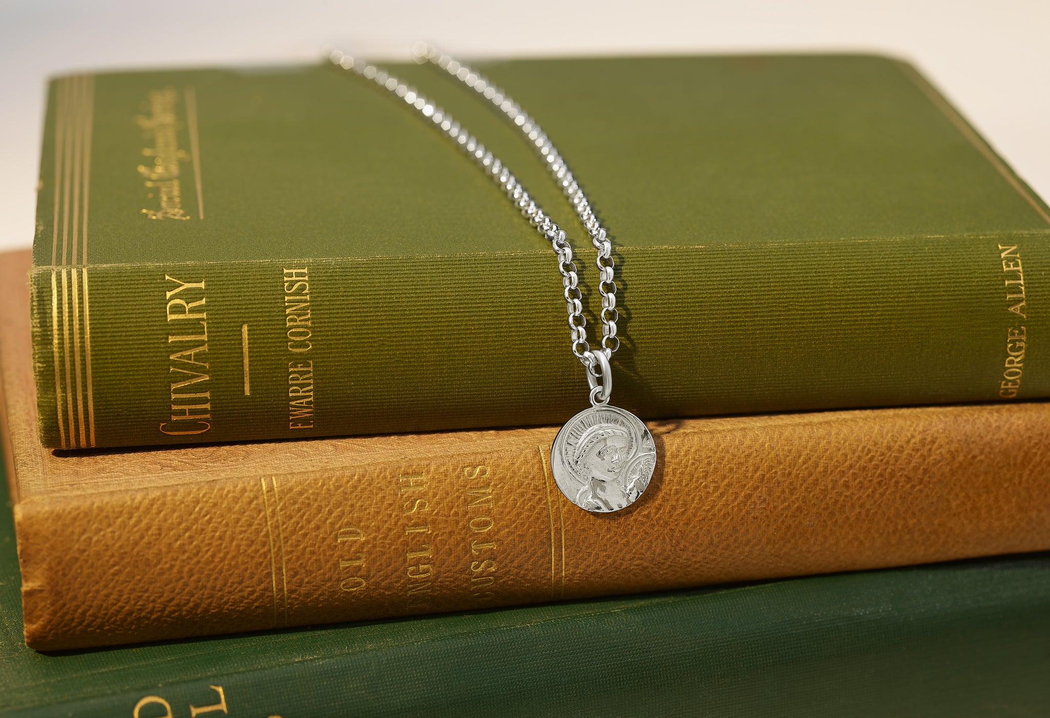 Handcrafted sterling silver round necklace pendant engraved with a saint and a chain necklace laid on some green books.